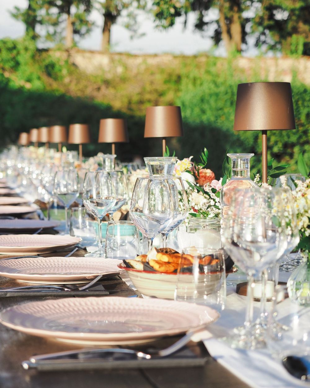 A long outdoor dinner table with glasses, flowers and lamps in a villa garden