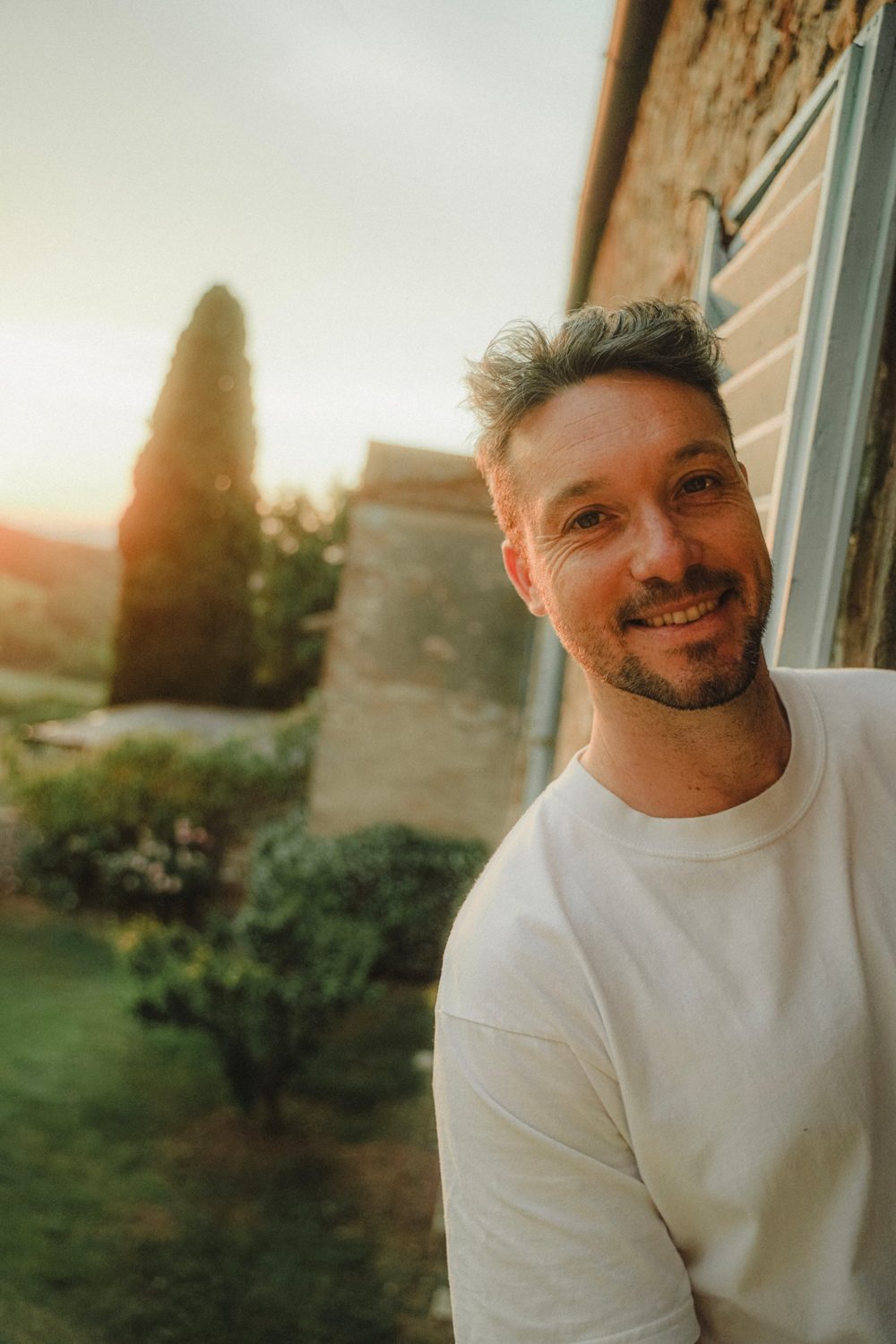Tom Scharre smiling outside a stone farmhouse at sunset, with a cypress tree behind him